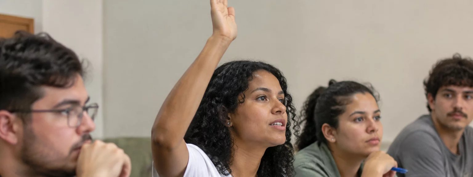 Participante com a mão erguida pedindo a vez, à mesa coberta de folhas escritas